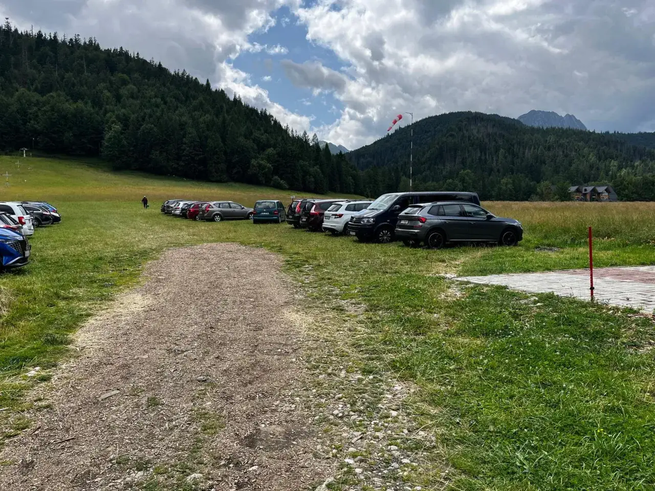 Samochody na trawiastym parkingu u podnóża gór. W oddali las i szczyty, widać wiatrowskaz. To parking przy Kasprowym Wierchu.