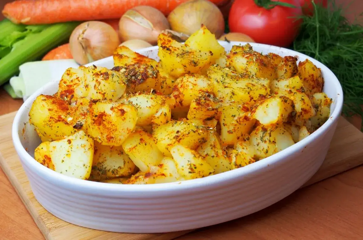 Pieczone ziemniaki w białej misce, posypane aromatyczną posypką do ziemniaków. W tle warzywa: marchew, cebula, pomidor i koperek.