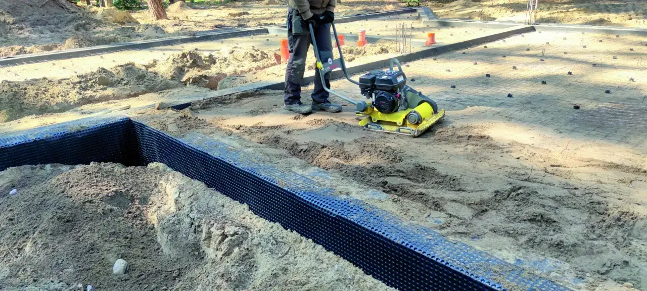 Pracownik zagęszcza ziemię zagęszczarką. Widać fundamenty, zbrojenie i czarną membranę. Chudziak ile cm?