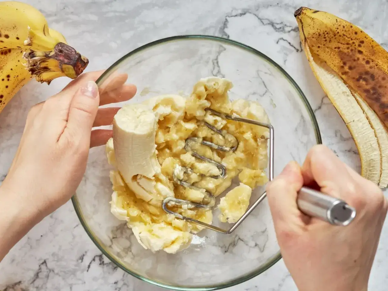 Ręce ugniatają banany w misce, przygotowując składniki na deser bananowy przepis. Obok leżą dojrzałe banany.