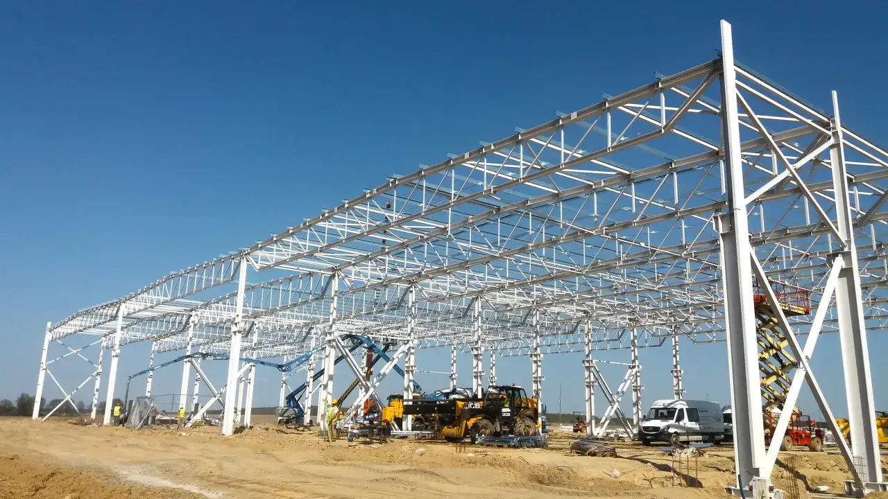 Trwa budowa hal przemysłowych. Widoczna stalowa konstrukcja, pracownicy na podnośnikach i maszyny budowlane na placu budowy.