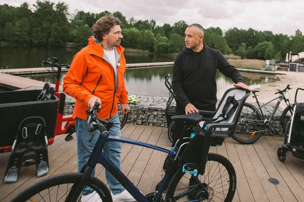 Dwóch mężczyzn rozmawia przy rowerach. Jeden z nich ma na sobie pomarańczową kurtkę, a drugi czarną bluzę. Obok nich stoi rower z zamontowanym fotelikiem na rower dla dziecka.