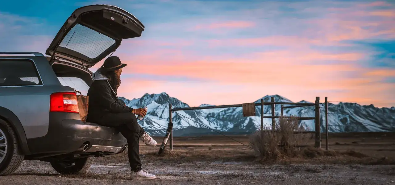 Osoba w kapeluszu siedzi na otwartym bagażniku samochodu, pijąc napój. W tle góry i zachód słońca. Idealne miejsce na spanie w samochodzie.
