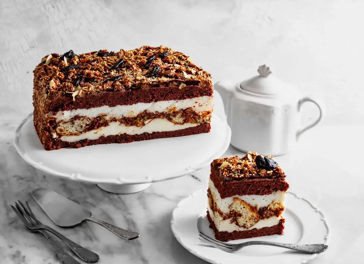 Przepis na ciasto kukułka bez pieczenia: czekoladowe warstwy przełożone kremem i biszkoptem, posypane orzechami i suszonymi owocami.