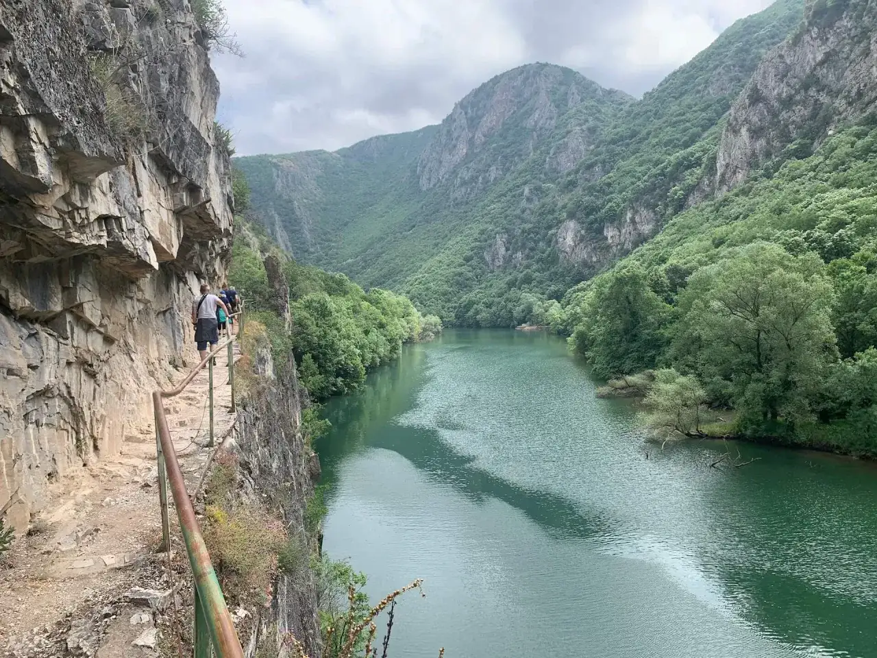 Spacerowicze wędrują wąską ścieżką wzdłuż skalnej ściany, podziwiając malownicze macedońskie g&oacute;ry i turkusową rzekę.