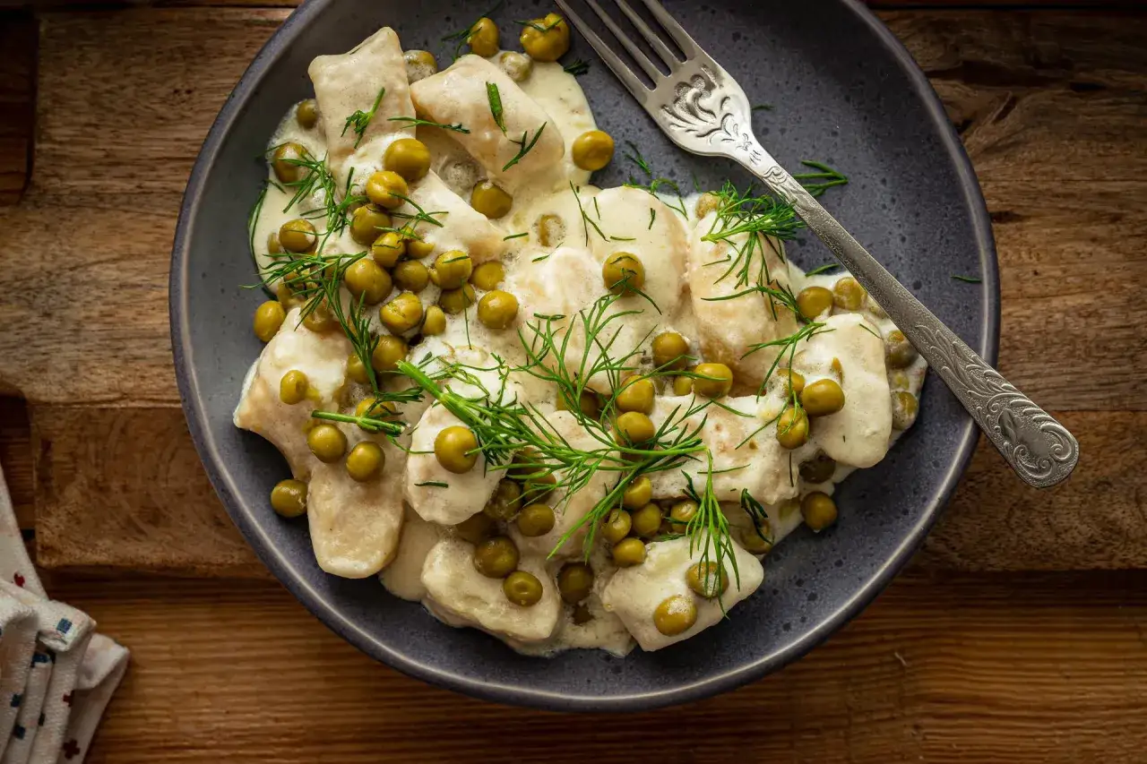 Pyszne kopytka w kremowym sosie z groszkiem i koperkiem. Idealne co do kopytek na obiad, podane na ciemnym talerzu z widelcem.