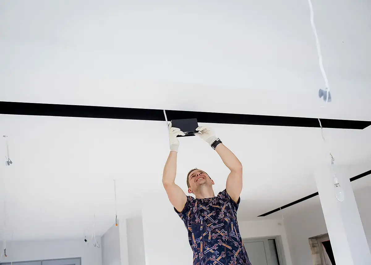 Mężczyzna w białych rękawiczkach montuje czarne pasy na suficie, tworząc nowoczesną instalację oświetleniową.
