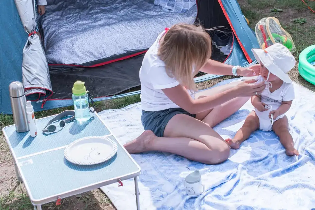 Mama przygotowuje dziecko do wyjścia z namiotu. Na stoliku termos, butelka wody, okulary. To lista co zabrać pod namiot z dziećmi.