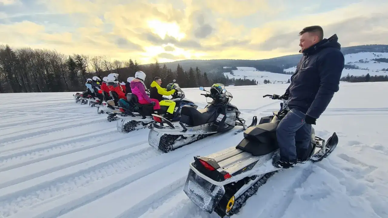 Grupa os&oacute;b na skuterach śnieżnych w Bukowinie czeka na rozpoczęcie zimowej przygody. Słońce przebija się przez chmury, oświetlając zaśnieżony krajobraz.