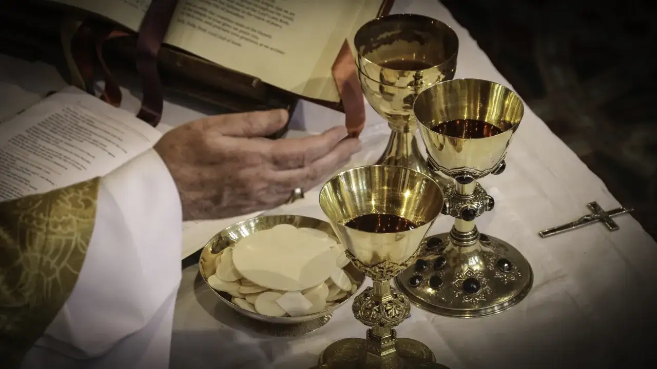 Dłoń księdza nad księgą, kielichy z winem i hostie. Tekst kanonu rzymskiego jest widoczny na otwartej księdze.