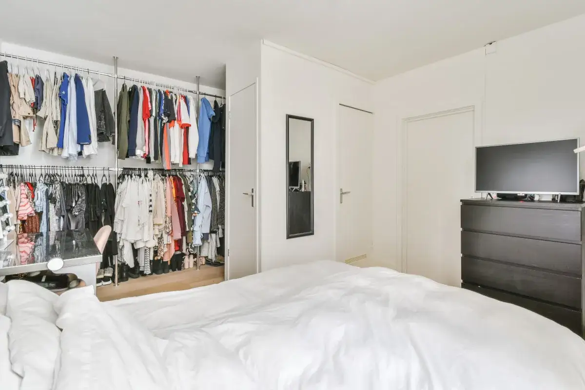 Otwarta garderoba w sypialni za ścianką, pełna ubrań na wieszakach. Obok łóżko z białym pościelą.