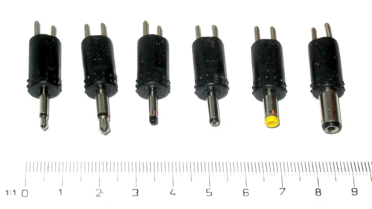 R&oacute;żne wtyczki elektryczne z metalowymi pinami i czarnymi obudowami, obok linijki.