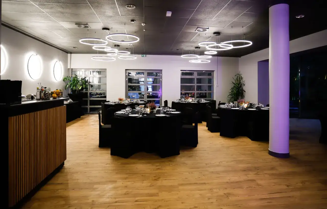 Elegancka sala bankietowa z okrągłymi stołami, przygotowana na imprezę dla 40 osób. Nowoczesne oświetlenie i drewniana podłoga tworzą przytulną atmosferę.