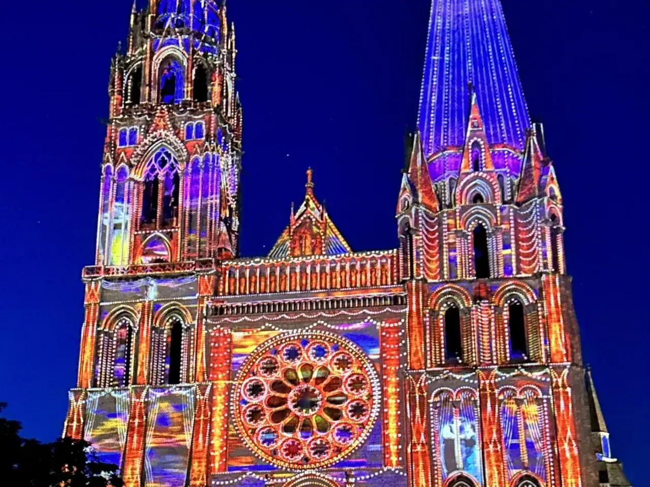 Katedra w Chartres nocą, rozświetlona kolorowymi projekcjami. Architektura gotycka w odcieniach niebieskiego i pomarańczowego.