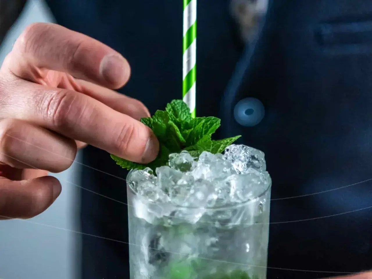 Dłoń dodaje listki mięty do orzeźwiającego mojito drink ze spritem, wypełnionego lodem i słomką.