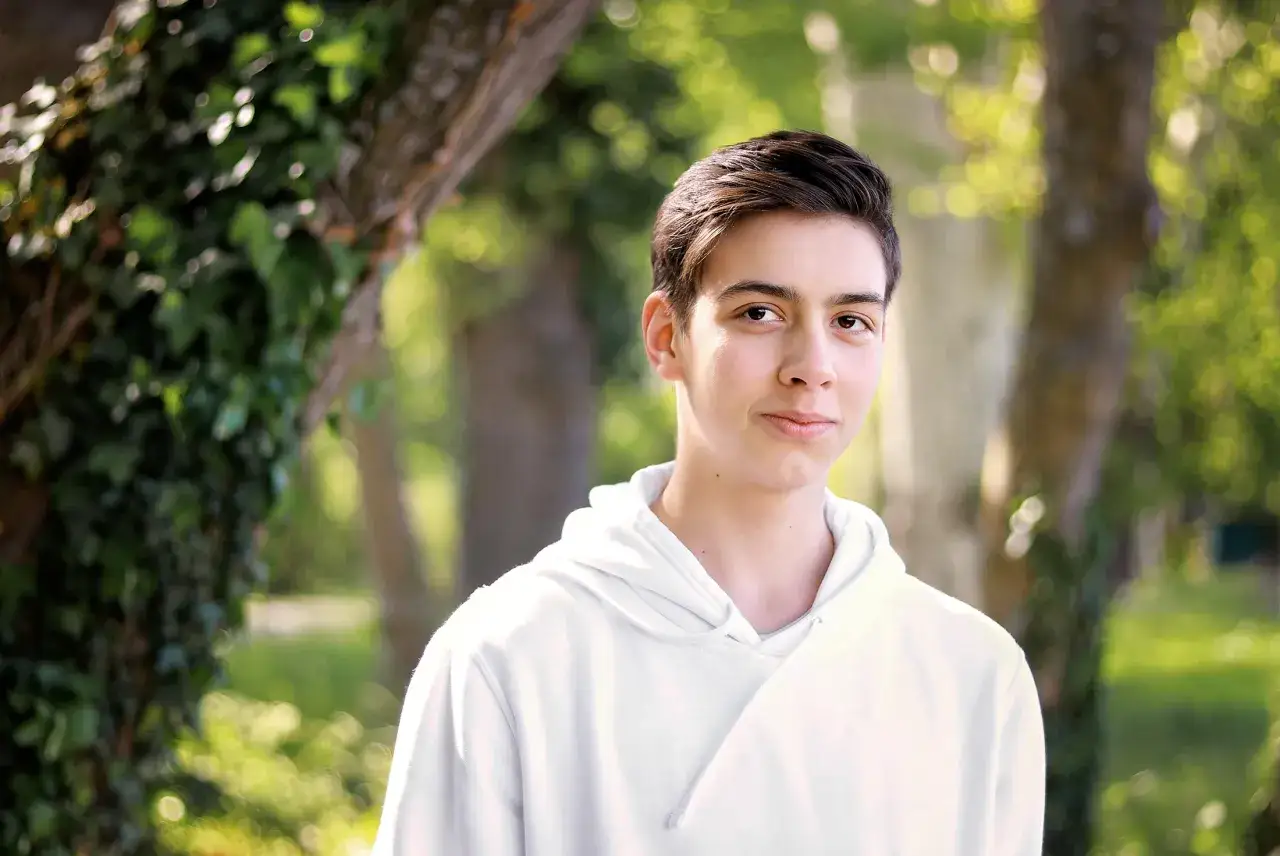 Chłopak w białej bluzie, uśmiechnięty, na tle zieleni. Piękny portret w plenerze.