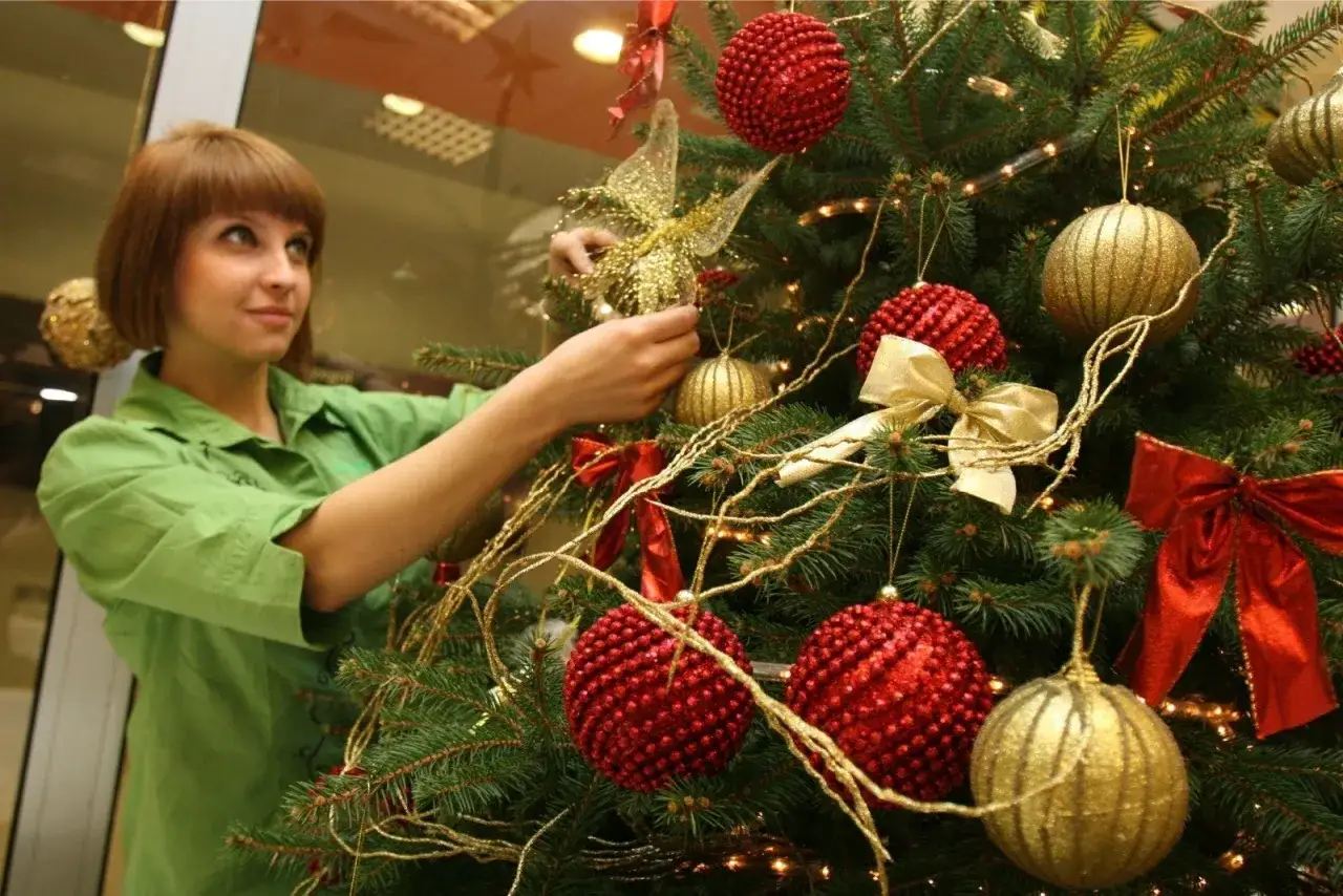 Kiedy zdjąć ozdoby świąteczne? Młoda kobieta w zielonej koszuli ozdabia choinkę złotymi i czerwonymi bombkami.