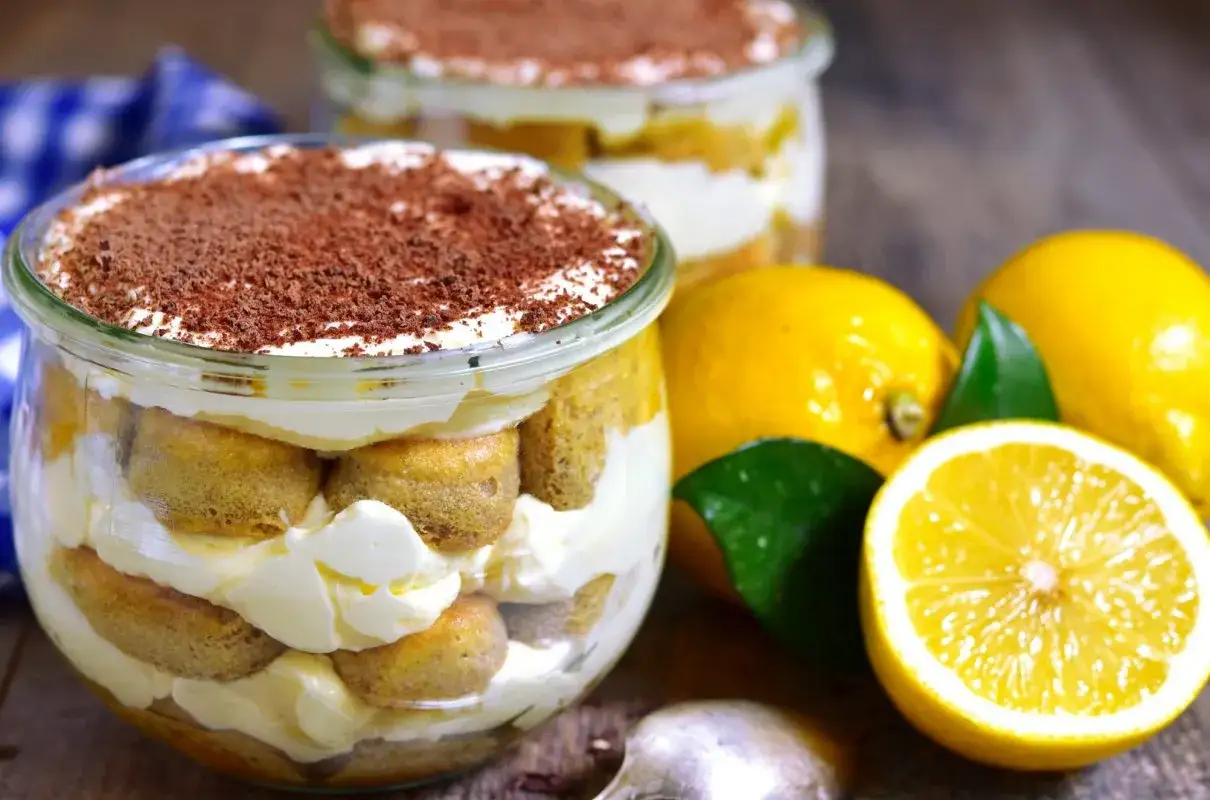 Pyszne tiramisu cytrynowe w słoiczku, przełożone biszkoptami i kremem, posypane czekoladą. Obok świeże cytryny.