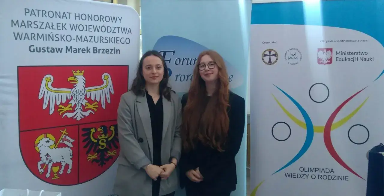 Dwie młode kobiety stoją przed banerem Olimpiady Wiedzy o Rodzinie.