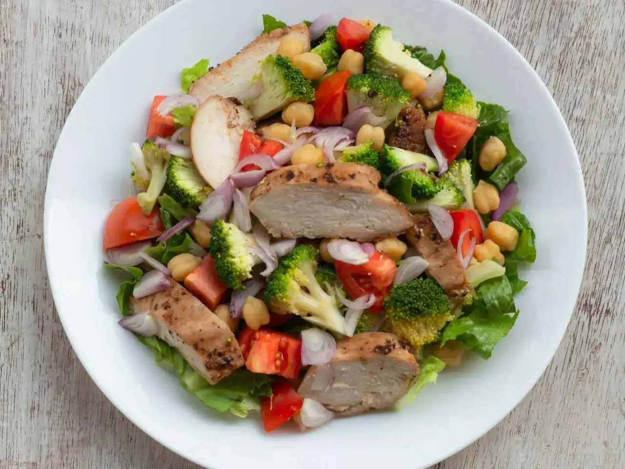 Pyszna sałatka z kurczakiem, brokułami, pomidorami i ciecierzycą na białym talerzu.
