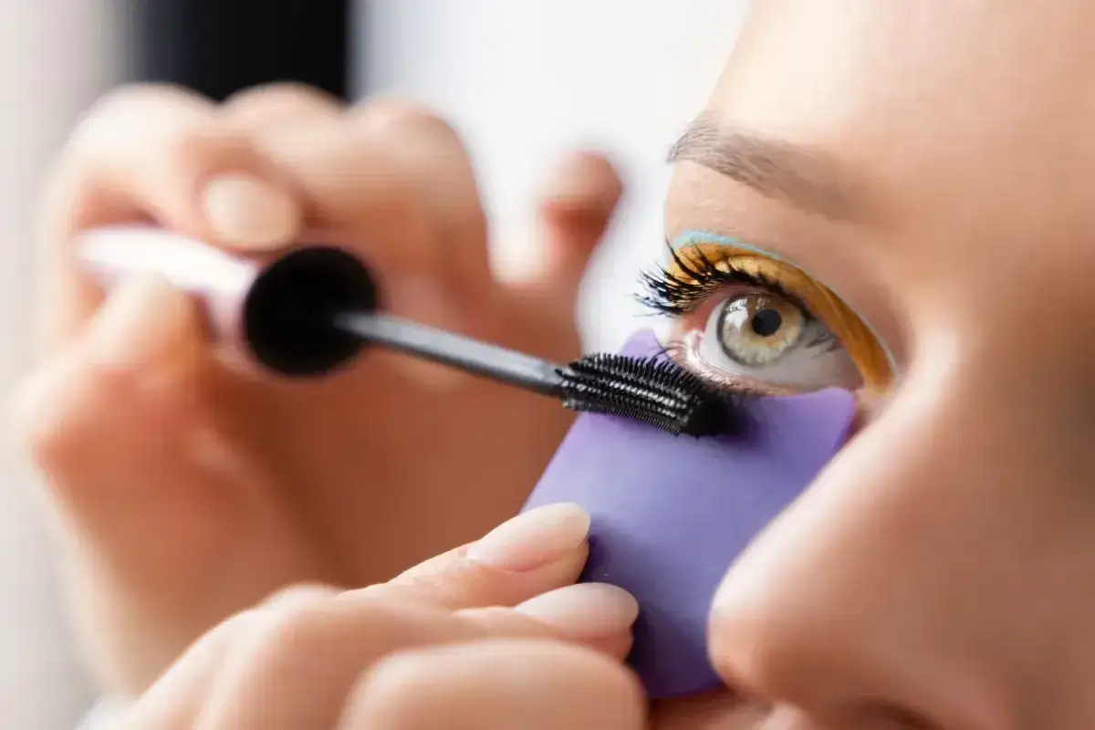 Osoba nakłada tusz na rzęsy, używając fioletowej osłonki. Czy można malować sztuczne rzęsy? Tak, ale te na zdjęciu są naturalne.