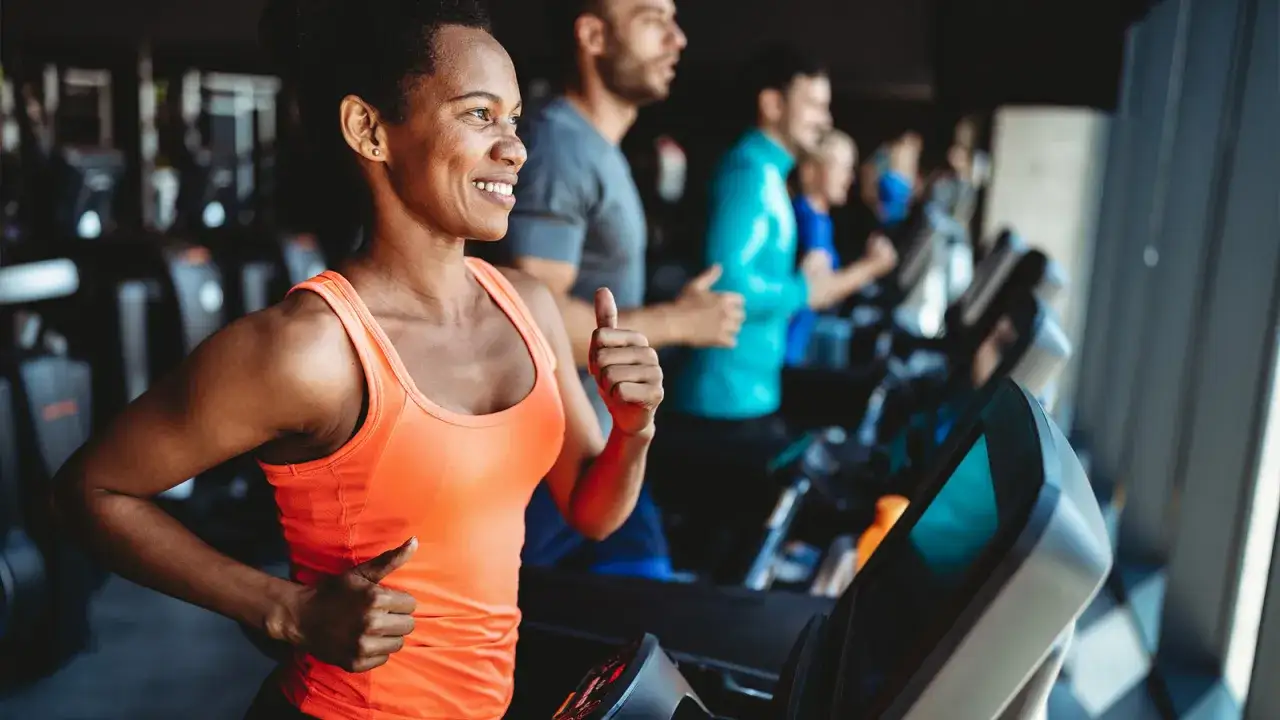 Uśmiechnięta kobieta w pomarańczowej koszulce daje kciuk w górę podczas treningu na bieżni. W tle inni ćwiczą.