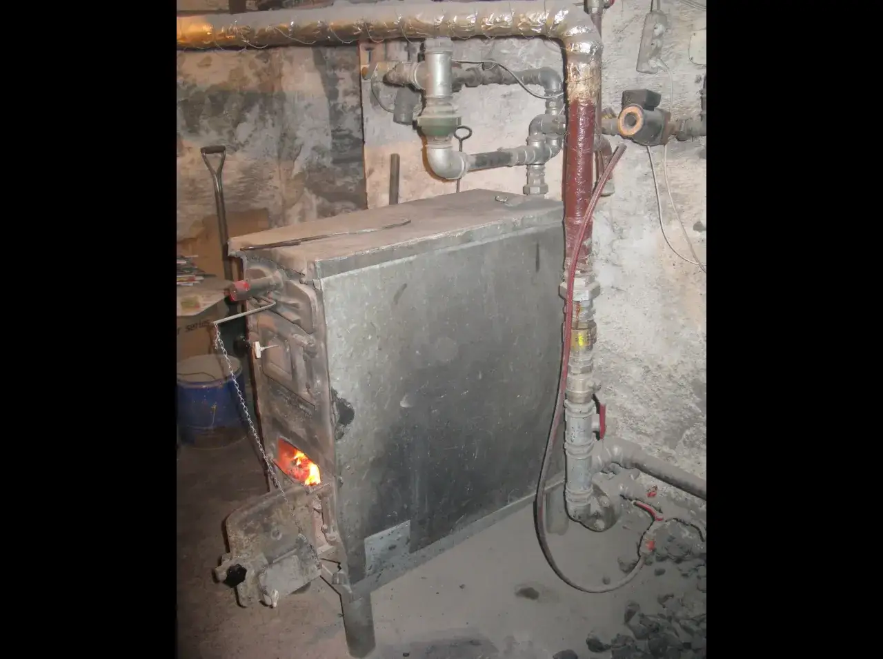 Stary piec węglowy z płomieniami w palenisku, otoczony rurami i narzędziami. Trudno ocenić, ile waży piec węglowy, ale jest solidny.