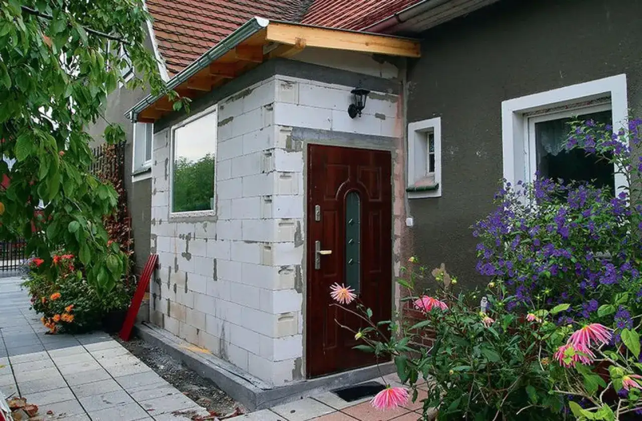 Rozbudowa domu na zgłoszenie: nowe ściany z bloczk&oacute;w, drzwi i okno czekają na wykończenie. Wok&oacute;ł kwitną kwiaty.