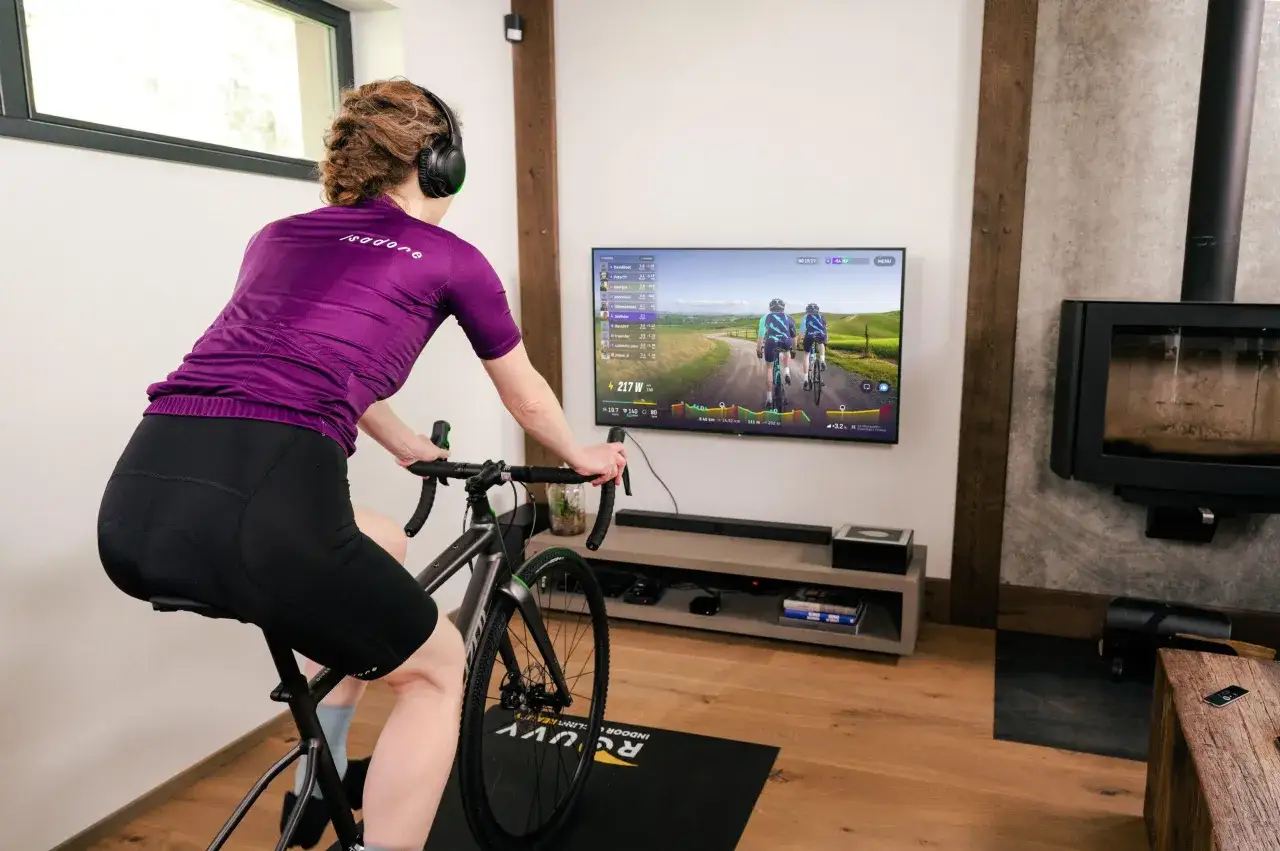 Kobieta na rowerku stacjonarnym, ćwicząca w domu, zastanawia się, czy rowerek stacjonarny odchudza brzuch, podczas wirtualnego wyścigu.