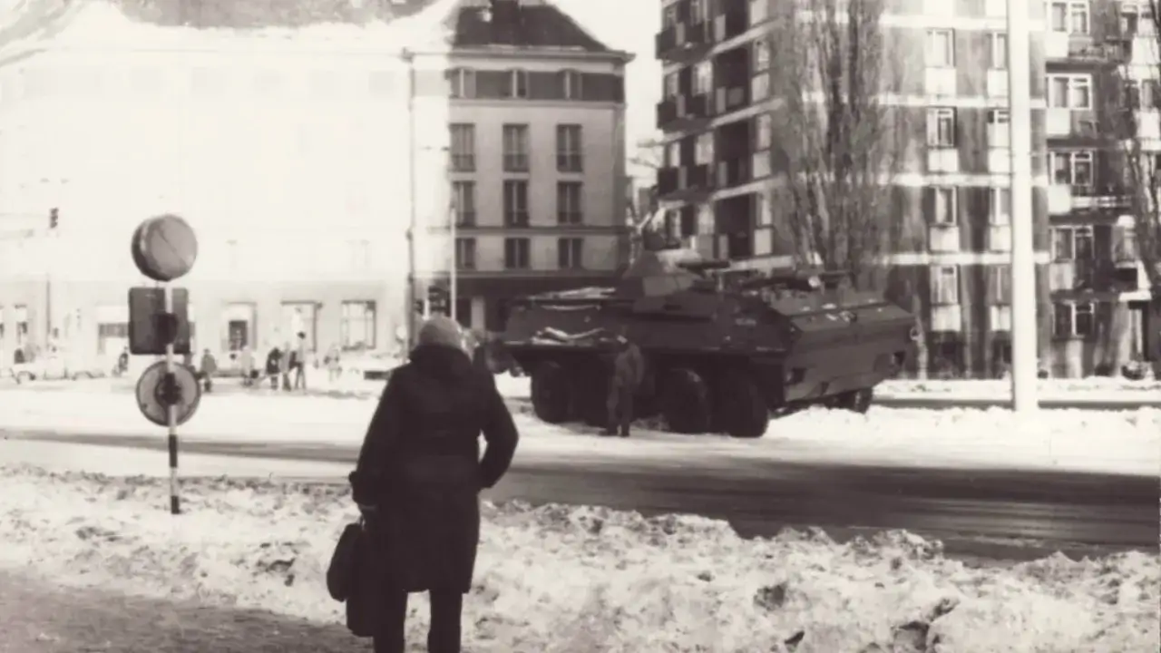 Zimowy dzień, kobieta z torbą idzie ulicą, obok pojazd opancerzony. To obraz z czas&oacute;w stanu wojennego.
