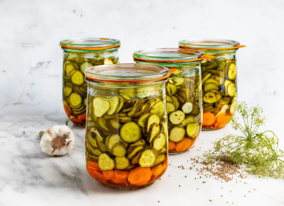 Słoiki z ogórkami i marchewką, gotowe do przygotowania sałatki szwedzkiej. Obok czosnek i koperek.