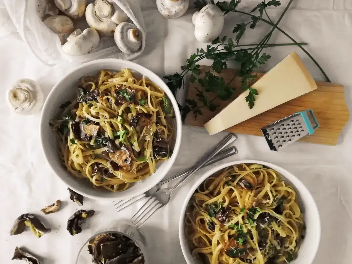 Dwa talerze makaronu z grzybami, posypane parmezanem i pietruszką. Obok świeże i suszone grzyby, kawałek parmezanu i tarka. Pyszny makaron z oliwą truflową.
