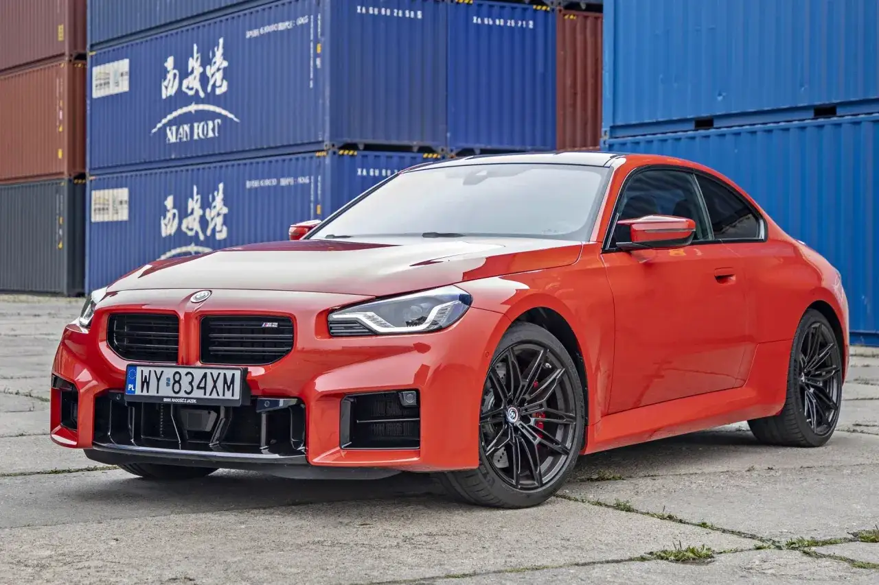 Pomarańczowe BMW M2 z czarnymi felgami, stojące na tle niebieskich kontenerów. Sprawdź aktualną bmw m2 cena!