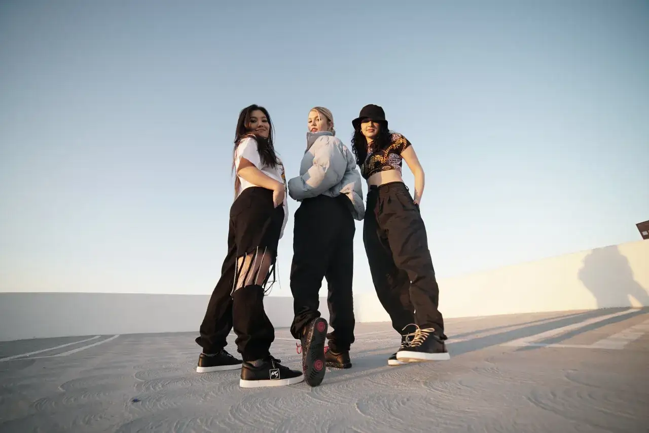 Trzy dziewczyny w stylowych ubraniach pozują na tle nieba, gotowe do zaprezentowania różnych rodzajów tańca.