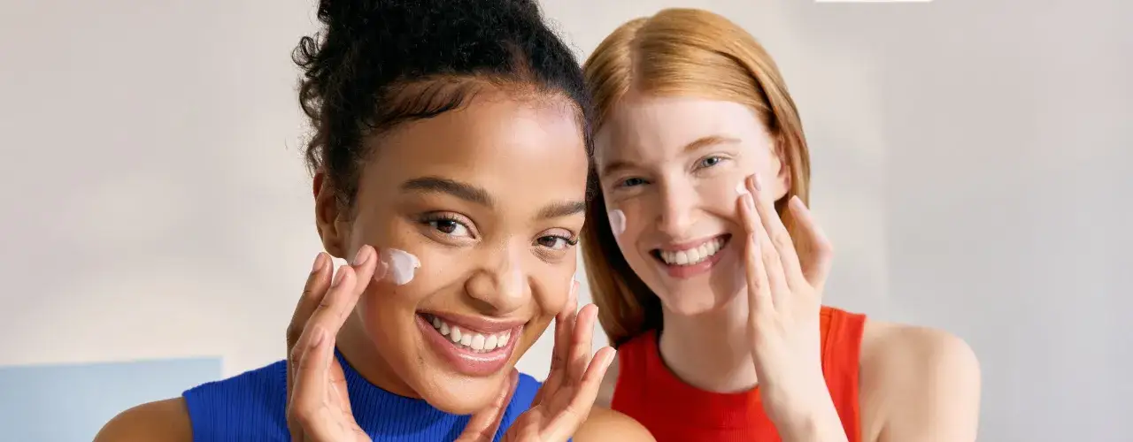 Dwie uśmiechnięte kobiety nakładają krem na twarz. Czy krem Nivea wysusza skórę? Te kobiety zdają się zaprzeczać, ich cera jest nawilżona i promienna.