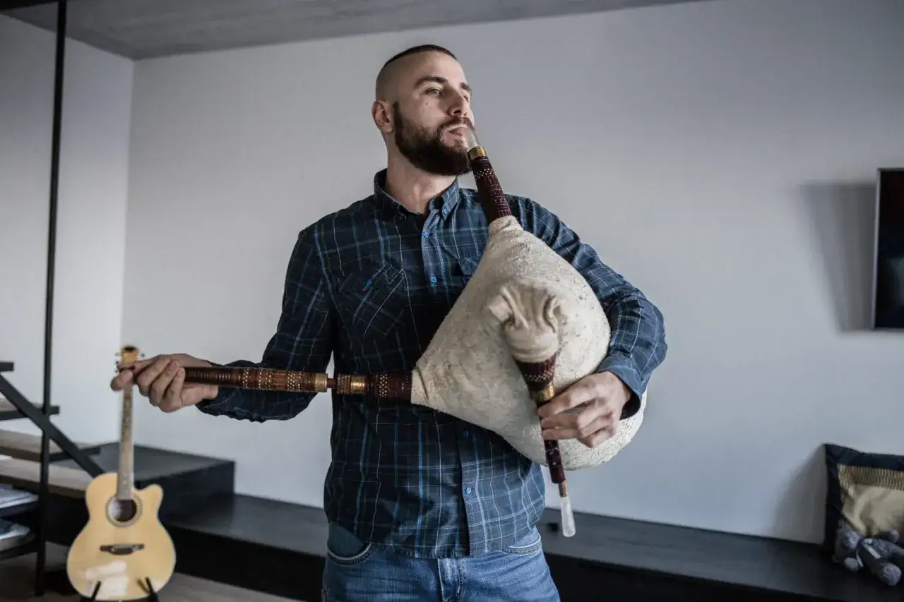 Mężczyzna z brodą, ludowy grajek z Ukrainy, dmie w dudy. Obok stoi gitara.