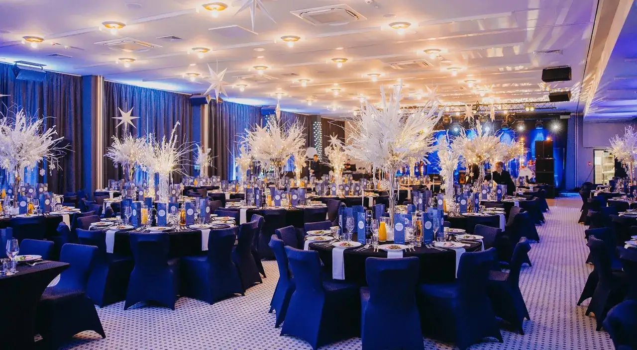 Elegancka sala bankietowa z niebieskimi obrusami i białymi dekoracjami, sugerująca zimowy motyw. Brak spójności między dress codem a wystrojem sali eventowej może być widoczny, gdy goście nie odzwierciedlają tej zimowej elegancji.