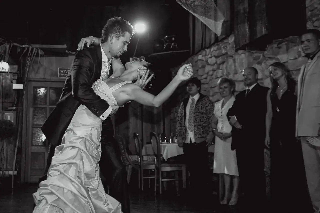 Para Młoda wykonuje pierwsze tańce weselne. Pan młody trzyma pannę młodą w romantycznym uścisku, podczas gdy goście obserwują ich z boku.