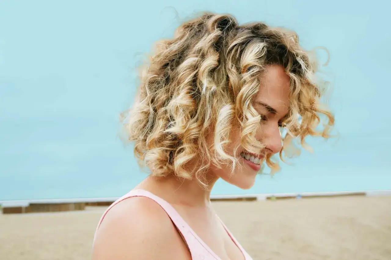 Uśmiechnięta dziewczyna z blond lokami, które wyglądają jak trwała na grube walki, na plaży.