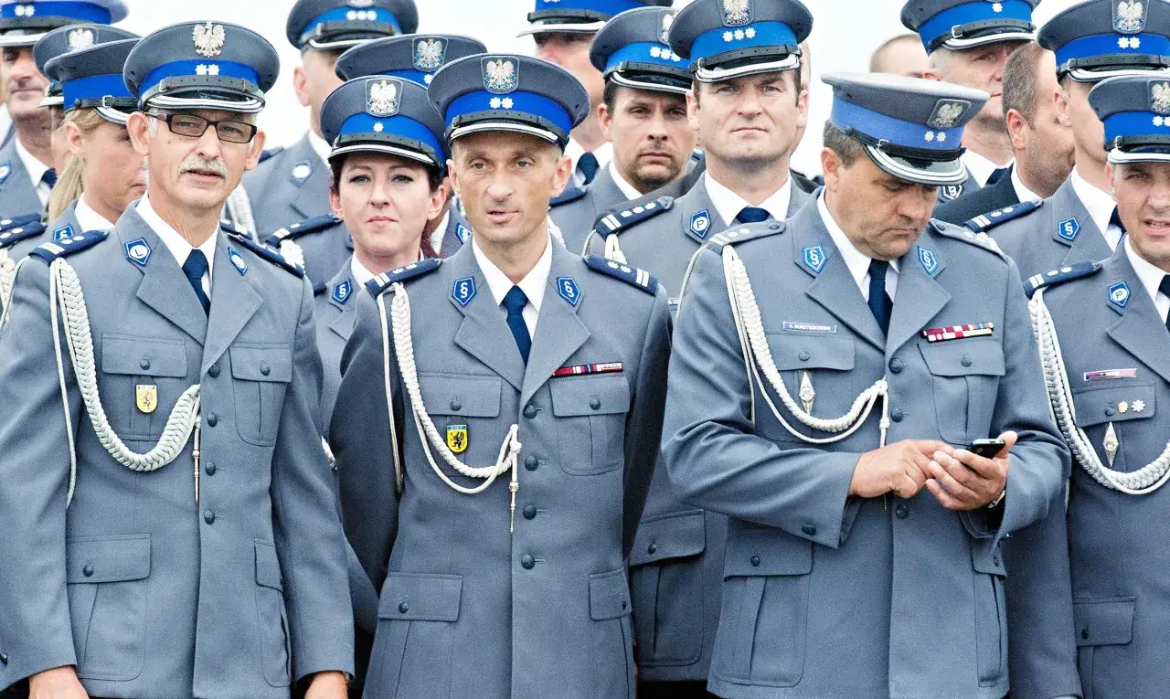 Policjanci w galowych mundurach, dyskutują w sprawie umundurowania policjantów. Jeden z nich sprawdza coś na telefonie.