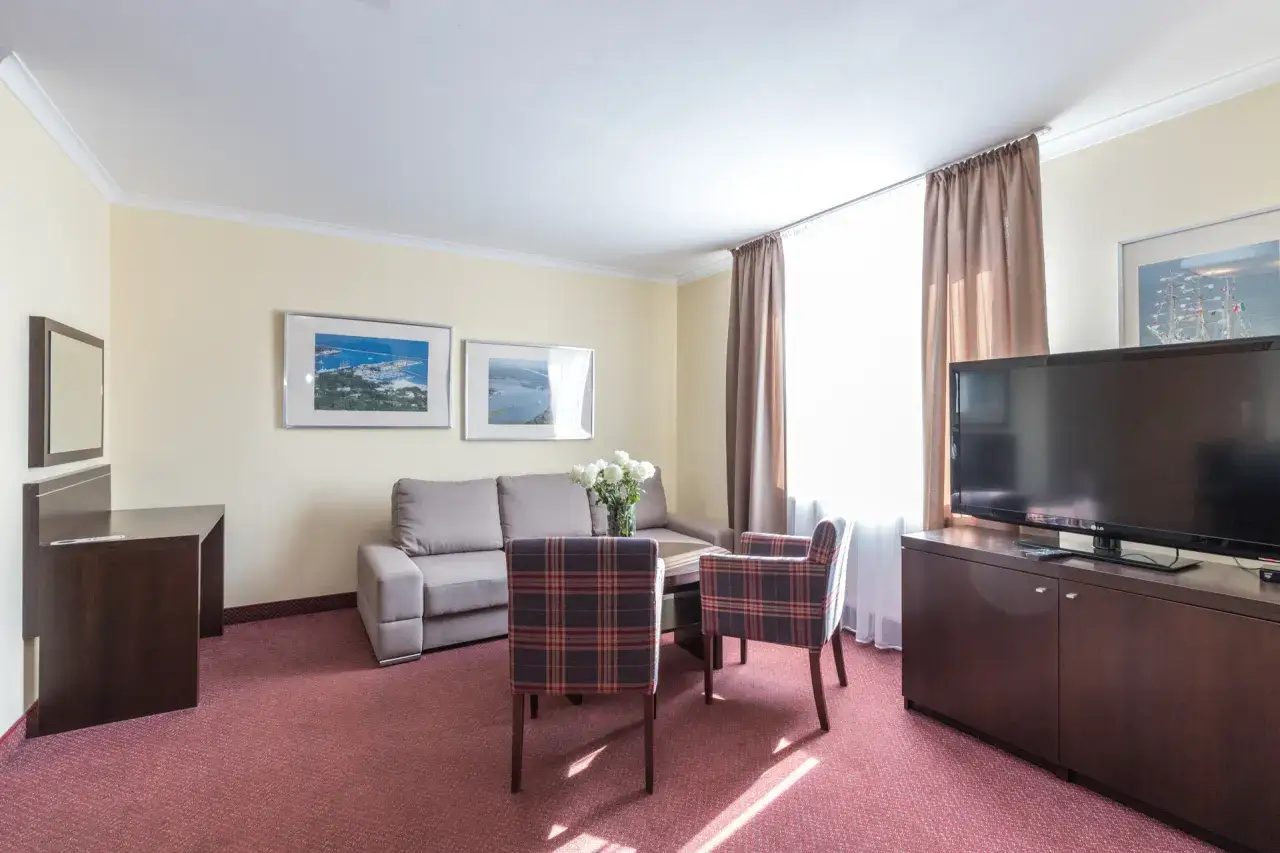 Przytulny pokój w resort spa plaza: kanapa, dwa fotele w kratę, stolik, biurko i telewizor.
