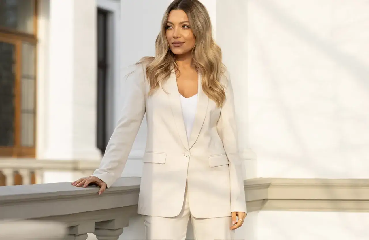 Elegancka kobieta w beżowym żakiecie i spodniach prezentuje stylowe biały żakiet stylizacje na tle klasycznej architektury.