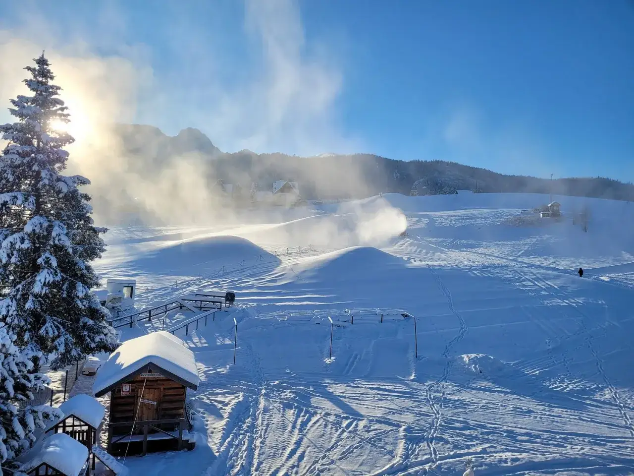Słońce przebija się przez mgłę nad stokiem narciarskim Budzowski Wierch. Działa armatka śnieżna, tworząc białą chmurę.