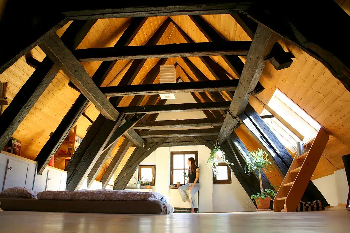 Przytulne poddasze z widoczną boazerią i drewnianymi belkami. Na dole ł&oacute;żko, przy oknie siedzi kobieta.