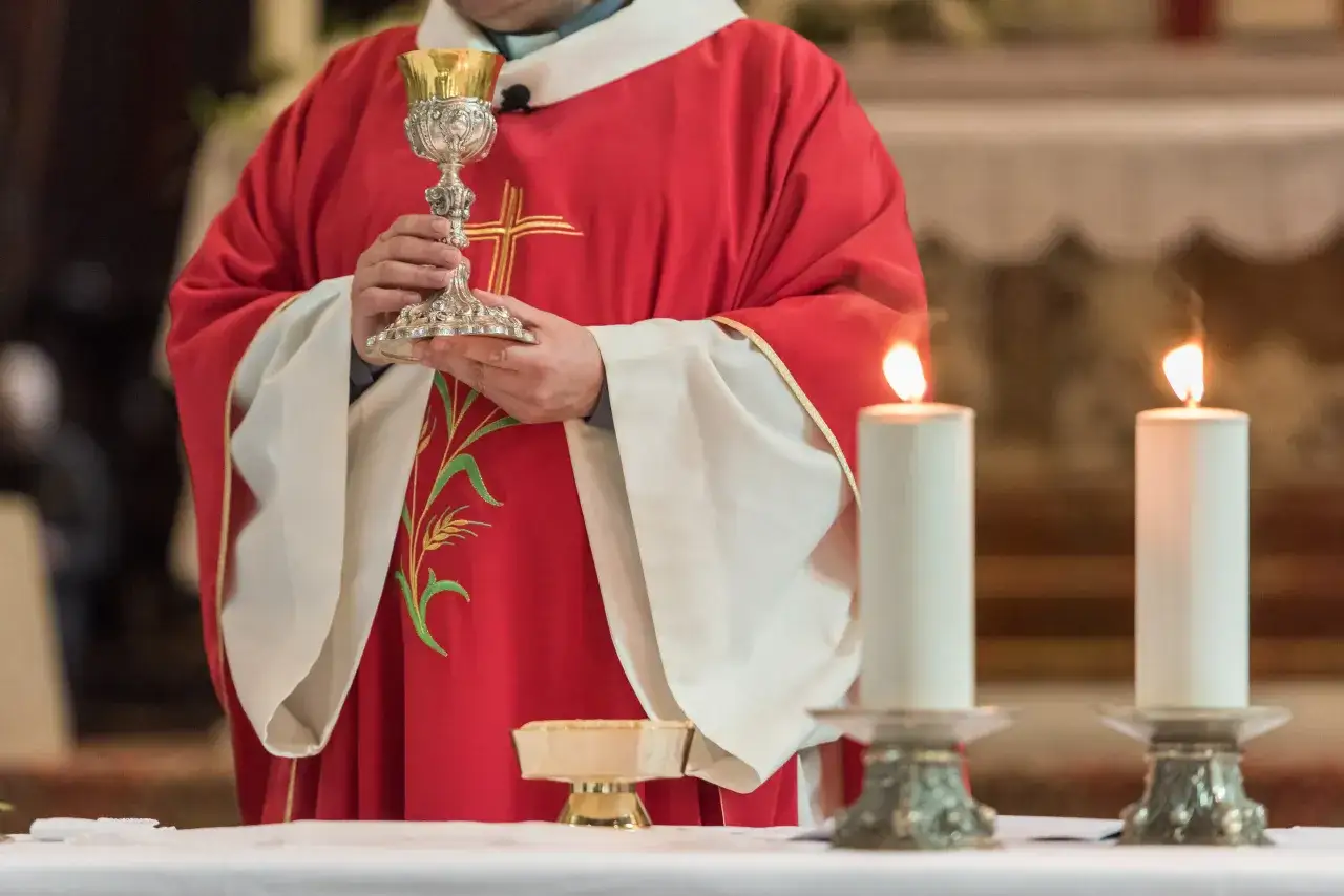 Kapłan w czerwonej szacie trzyma kielich. Obok płoną świece. To fragment uroczystości, która może być związana z konsekracją kościoła.