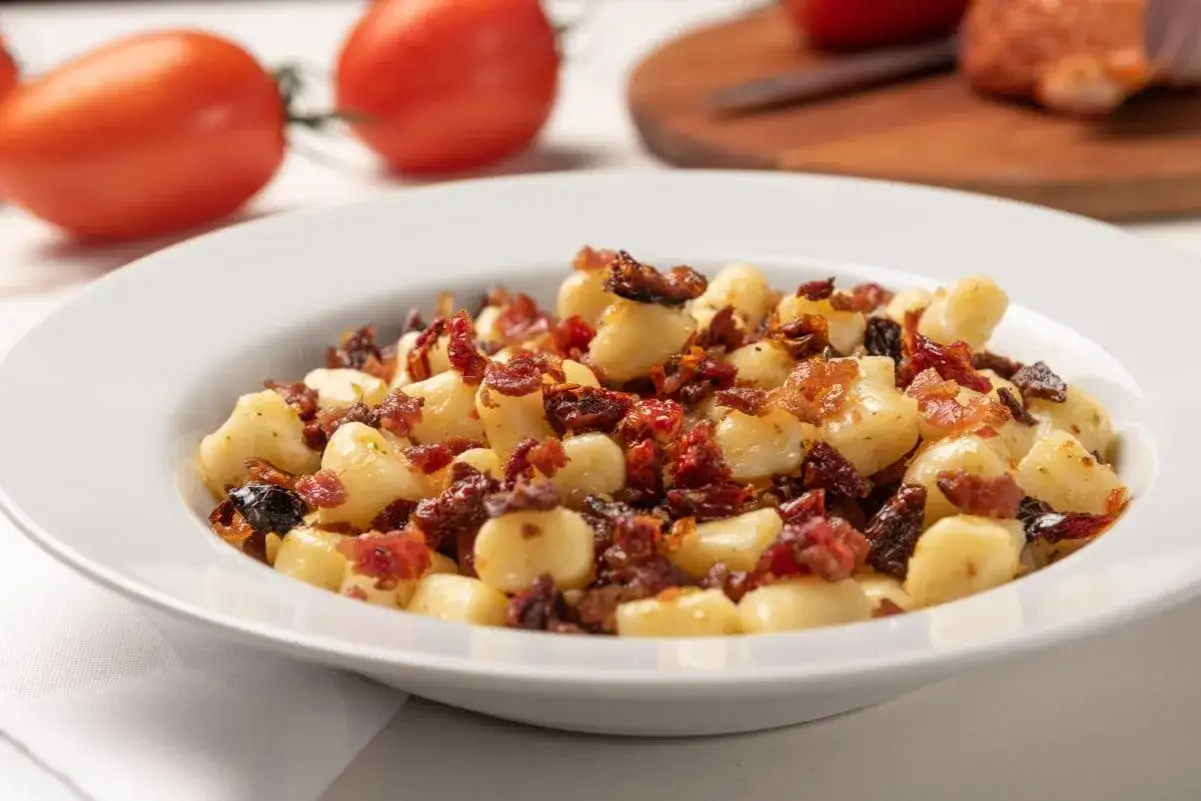 Pyszne gnocchi z boczkiem i suszonymi pomidorami na białym talerzu, w tle pomidory.