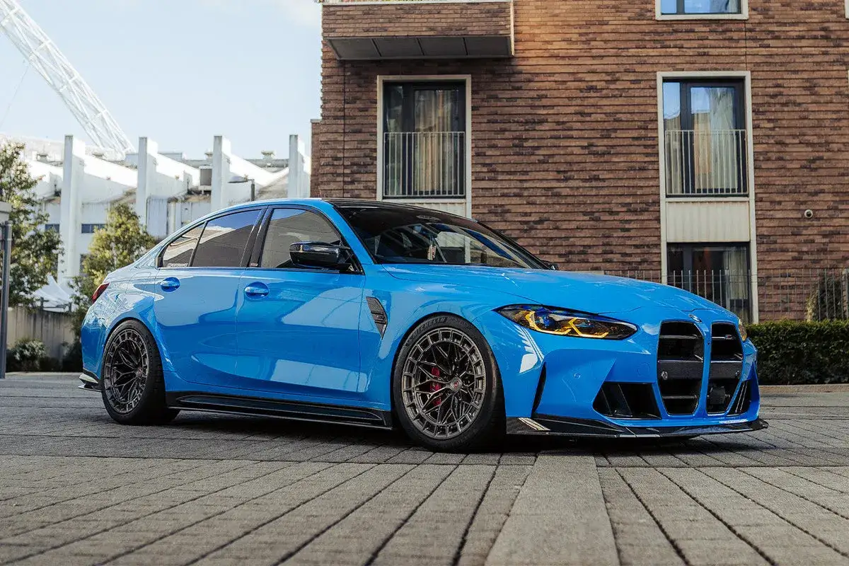 Błękitne BMW M3 G80 z przyciemnianymi szybami i sportowym pakietem aerodynamicznym stoi na brukowanej nawierzchni przed nowoczesnym budynkiem.