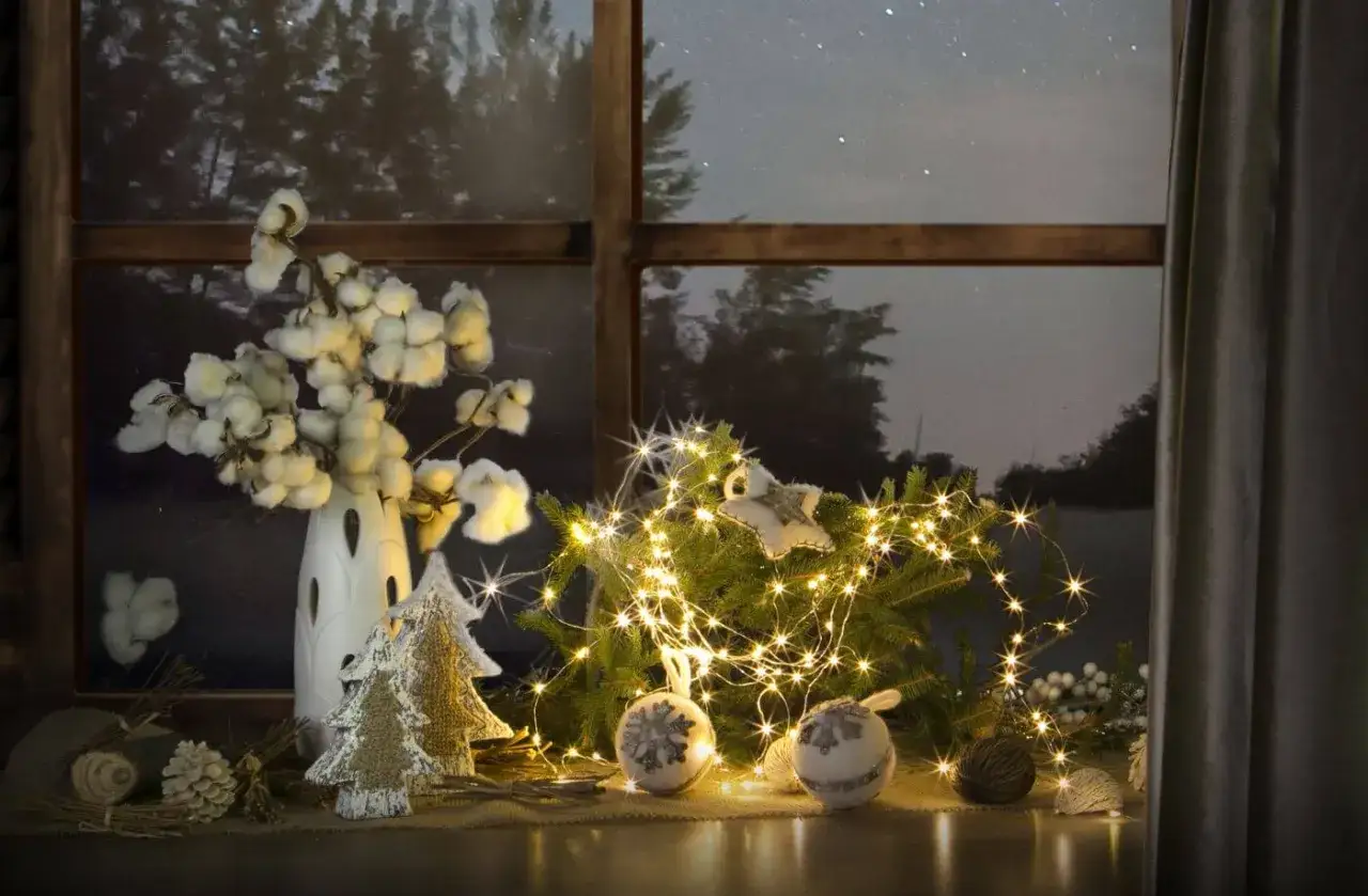 Świąteczne dekoracje okien: ozdoby na parapet z gałązkami bawełny, choinką i lampkami tworzą przytulny nastr&oacute;j.