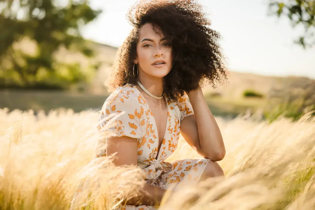 Kobieta z burzą lok&oacute;w w kwiecistej sukience siedzi w trawie. Idealne miejsce do robienia zdjęć w złocistym świetle.