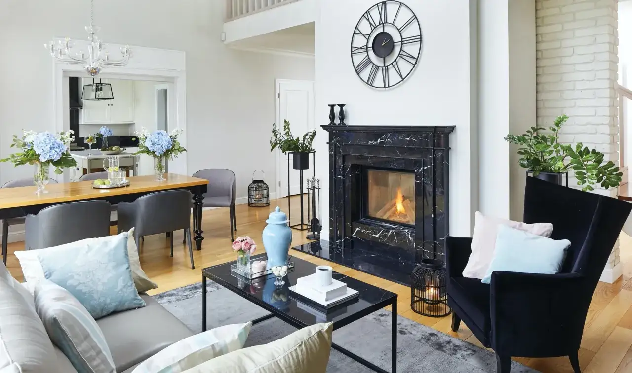 Przytulny kominek w salonie z palącym się ogniem, czarnym marmurowym obramowaniem i stylowym zegarem na ścianie.